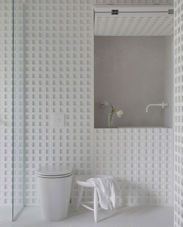 Detalle lavamanos de baño de habitación e inodoro, con grifería en blanco, lavamanos en hueco abierto junto a jarrón con flores blancas y estructura perforada con motivos geométricos