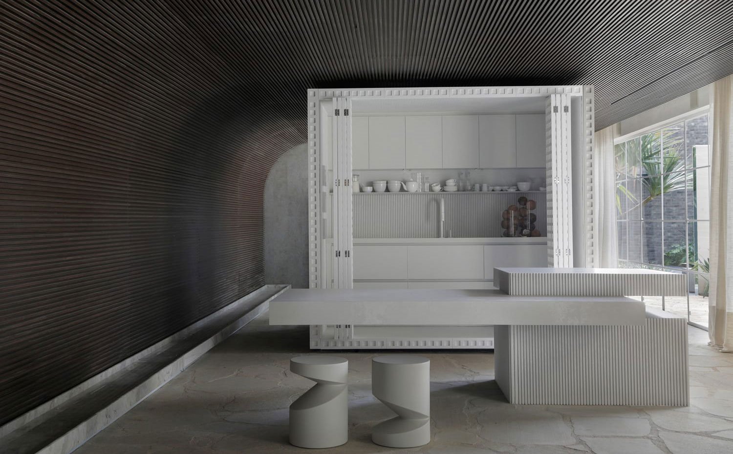 Módulo blanco con cocina integrada y puertas plegables, isla de trabajo con base blanca alistonada y encimera volada para office, dos taburetes blancos
