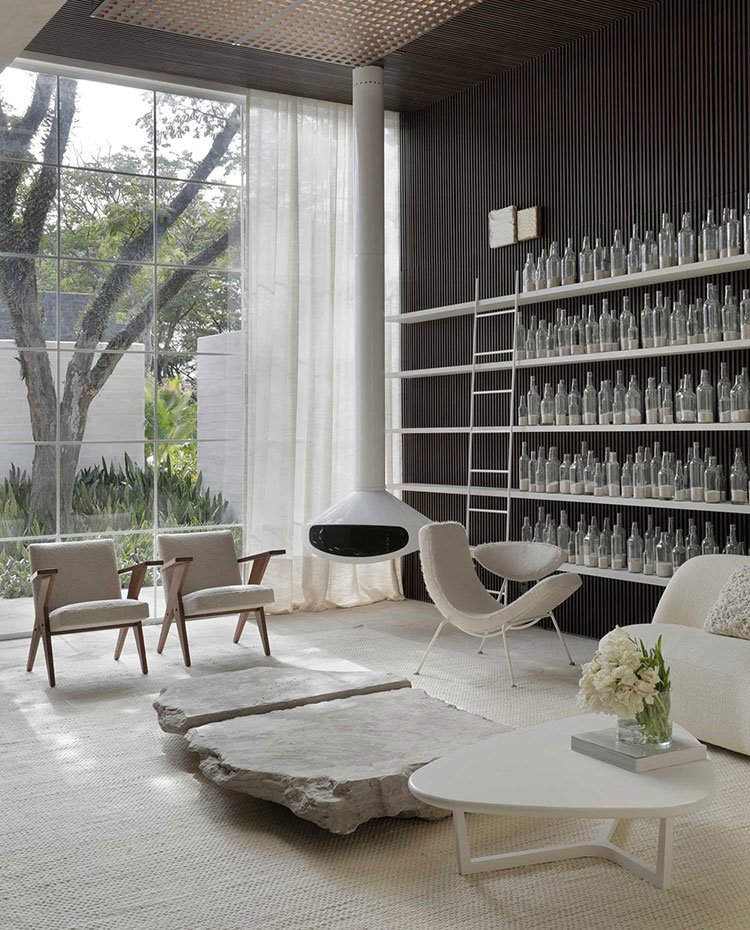 Salón con estantería de botellas de cristal, chimenea suspendida, butacas, sofá y mesa de centro en blanco, cerramiento acristalado con cortina blanca de techo a suelo