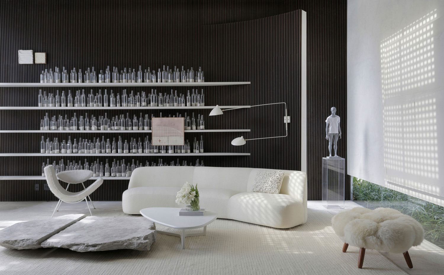 Zona de estar con mobiliario en blanco, estantería con botellas de cristal, escultura en gris sobre pedestal de metacrilato, pared alistonada
